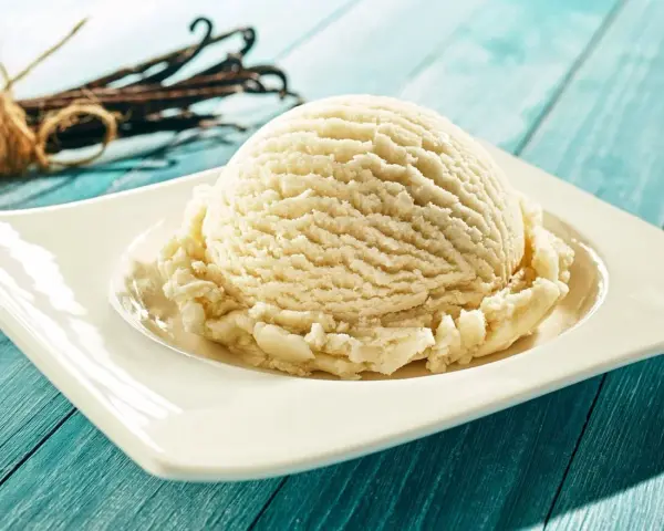 Une boule de glace maison à la vanille, servie sur une assiette blanche, avec des gousses de vanille en arrière-plan.