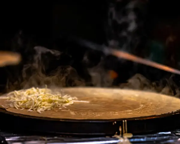 Fromage râpé fondant sur une crêpe chaude, vapeur s'élevant. Une tâche de nettoyage crêpière Tefal brûlée est visible sur le bord.