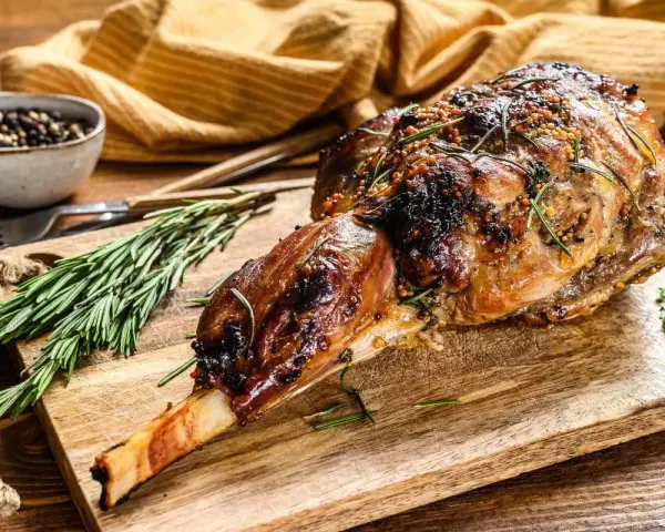 Selle d'agneau rôtie, garnie de romarin et de moutarde, sur une planche en bois avec des herbes fraîches.