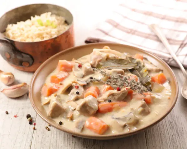 Un plat de blanquette de veau crémeuse avec des carottes, des champignons et des herbes, servi avec du riz dans une casserole en cuivre.