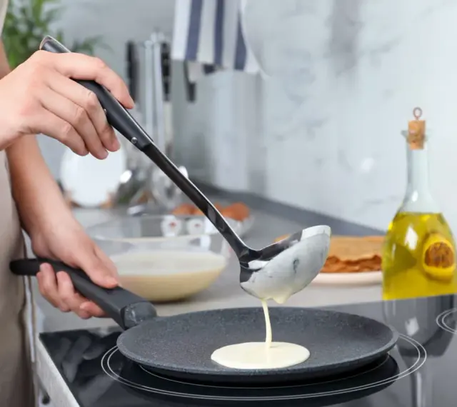 Une louche verse de la pâte dans une poêle pour une recette pâte à crêpe facile. Huile d'olive et pile de crêpes en arrière-plan.