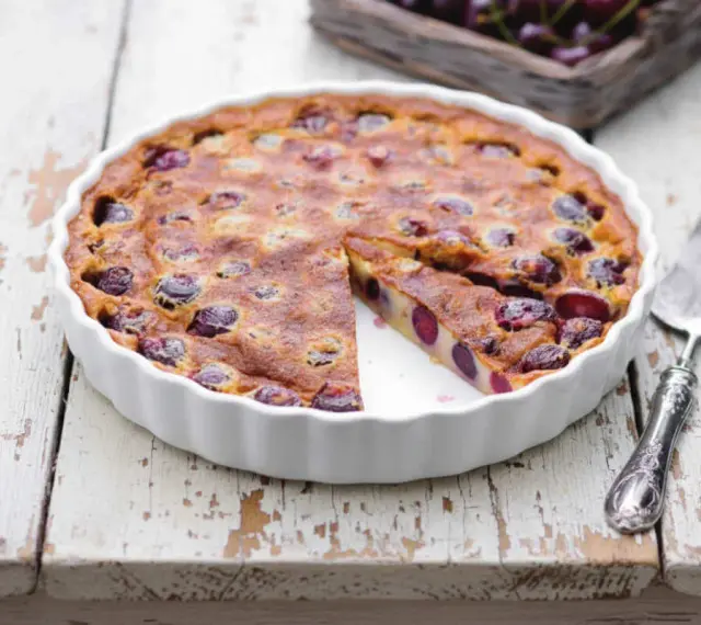 Un délicieux clafoutis aux cerises, encore chaud dans son plat blanc, avec une part coupée.