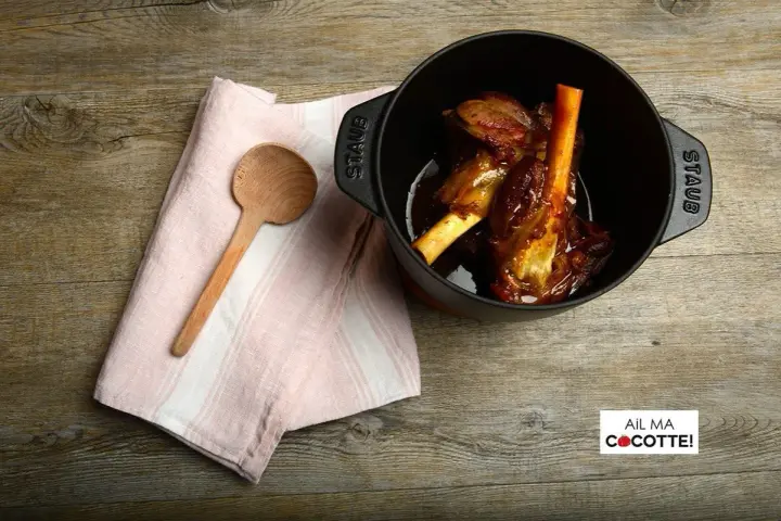 Souris d'agneau à la cocotte en fonte, tendres et savoureuses, prêtes à être dégustées. Une cuillère en bois et une serviette rose complètent la scène.