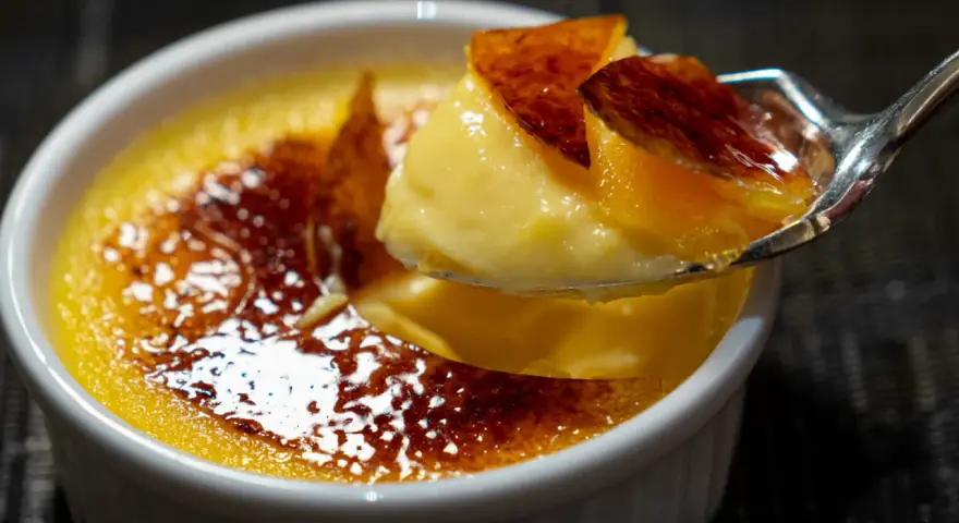 Une cuillère prélève un morceau de crème brûlée, révélant sa texture onctueuse sous une croûte de caramel craquante.