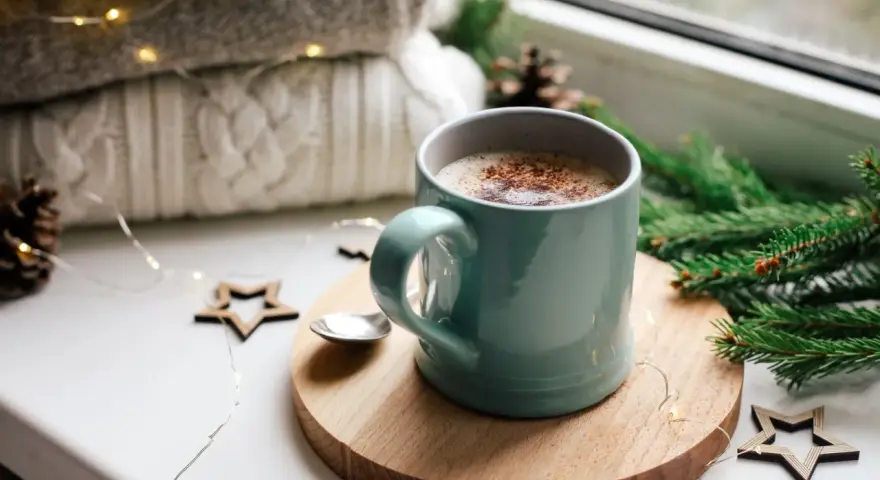 Une tasse de chocolat chaud fumant, entourée de décorations de Noël et de lumières scintillantes, promet un moment de réconfort.