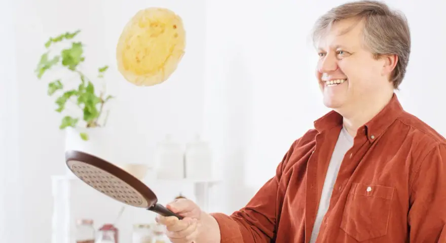 Un homme souriant s'amuse à retourner une crêpe en plein vol avec une poêle.