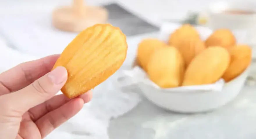 Une main tient une madeleine dorée, parfaite pour une recette madeleine moelleuse. Un bol rempli de ces délicieuses pâtisseries est visible en arrière-plan.