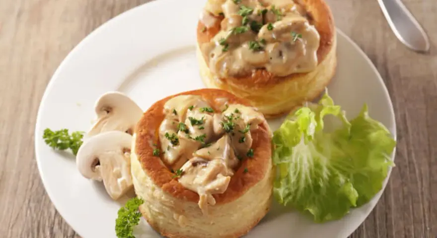 Deux bouchées à la reine garnies d'une sauce crémeuse aux champignons, accompagnées de champignons frais et d'une feuille de laitue.