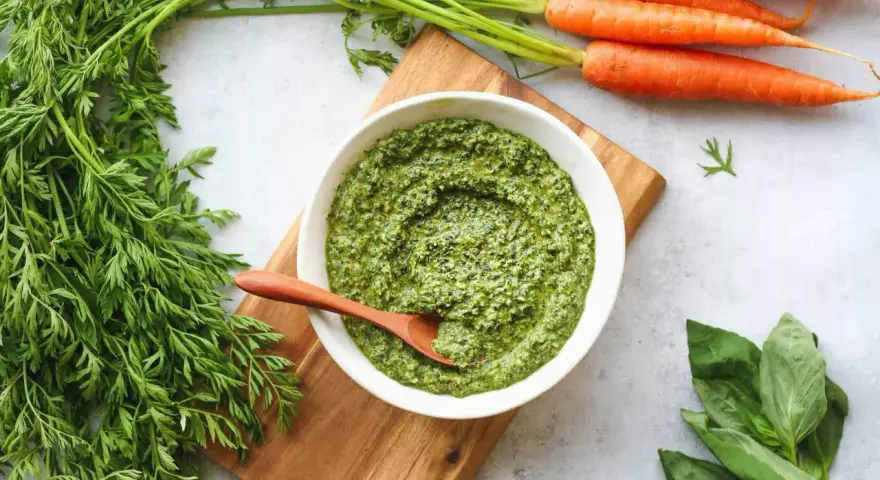 Un bol de pesto de fanes de carottes, avec des carottes fraîches et du basilic à côté.
