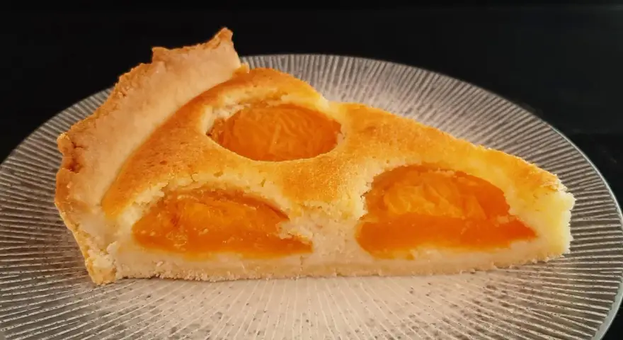 Une part de tarte aux abricots dorée, avec des fruits juteux incrustés dans une pâte moelleuse, sur une assiette texturée.