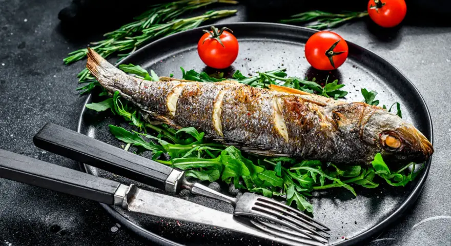 Un bar entier, parfaitement doré, est prêt pour la dégustation. Ce plat de cuisson bar au four est servi sur un lit de roquette fraîche, accompagné de tomates cerises et de romarin.