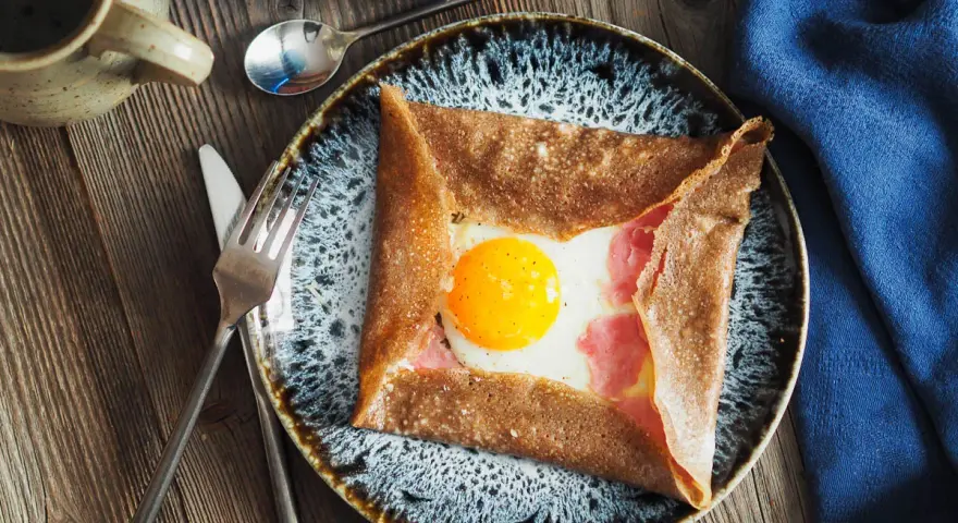 Une galette de sarrasin garnie d'un œuf au plat et de jambon, un des nombreux types de crêpes salées. Servi sur une assiette avec des couverts.