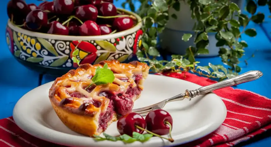 Une part de tarte aux cerises, garnie de menthe fraîche, sur une assiette blanche avec une fourchette. Un bol de cerises est visible en arrière-plan.