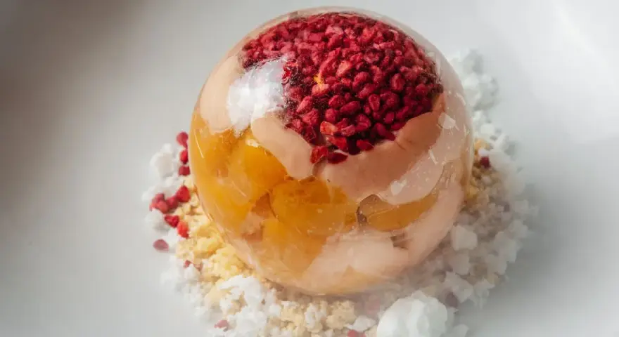 Sphère transparente garnie de fruits rouges et de pêche melba, sur un lit de miettes et de glace pilée.