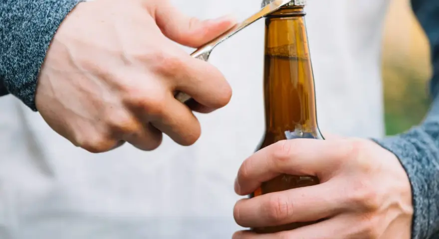Main gauche tenant une bouteille, main droite utilisant un objet pour ouvrir la bouteille sans tire bouchon.