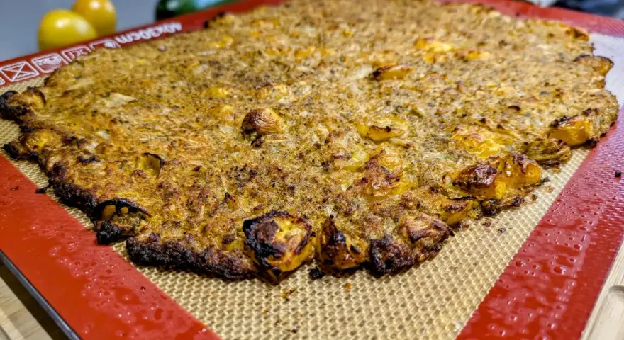 Une galette complète au four, dorée et croustillante, sur un tapis de cuisson en silicone. Des tomates jaunes et une courgette sont floues en arrière-plan.