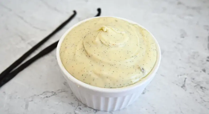 Un bol de ganache chocolat blanc onctueuse, parsemée de points de vanille, avec deux gousses de vanille à côté.