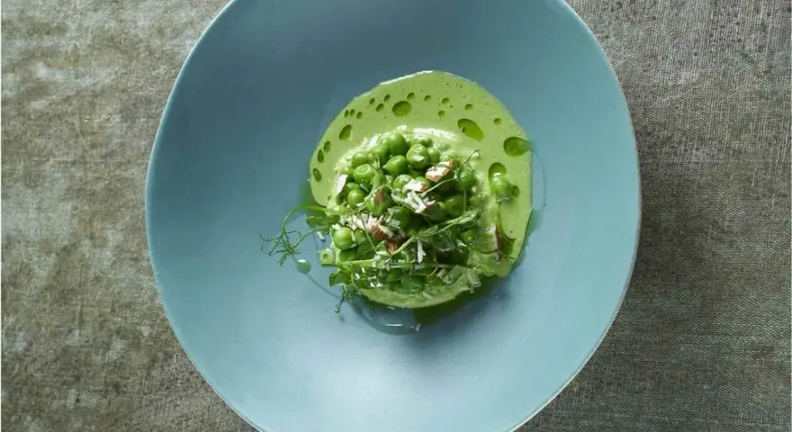 Délicieuse cuisson petit pois frais, servis dans une assiette bleue avec une émulsion verte et des éclats d'amandes.