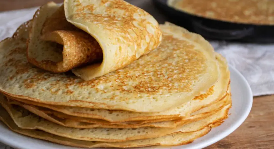 Pile de crêpes dorées, une roulée sur le dessus. Une poêle avec une crêpe en cuisson est visible en arrière-plan. Parfait pour 1 litre de pâte à crêpe.
