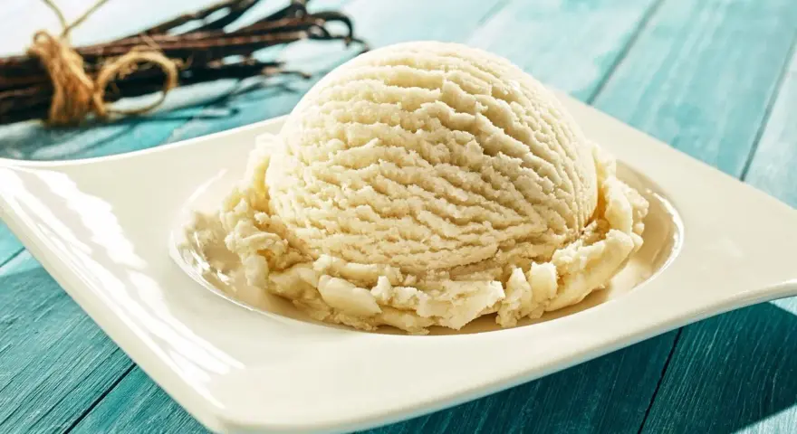 Une boule de glace maison à la vanille, servie sur une assiette blanche, avec des gousses de vanille en arrière-plan.