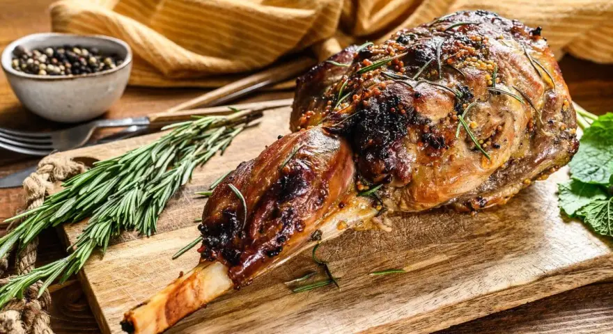 Selle d'agneau rôtie, garnie de romarin et de moutarde, sur une planche en bois avec des herbes fraîches.