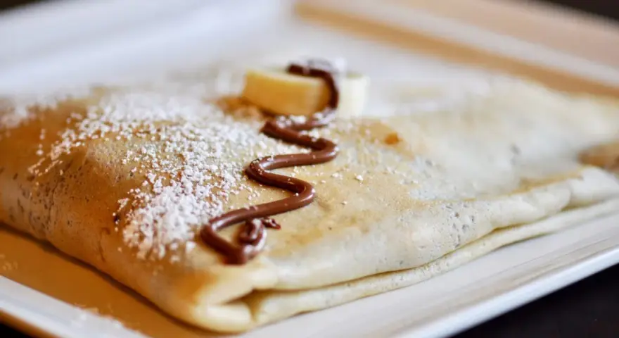 Crêpe pliée, saupoudrée de sucre glace, décorée de chocolat fondu et d'une rondelle de banane. La meilleure recette de pâte à crêpes pour un délice !