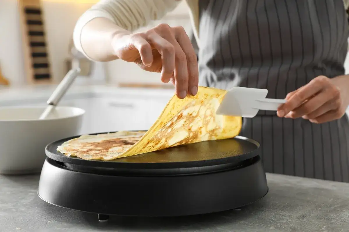 Une main soul&egrave;ve une cr&ecirc;pe dor&eacute;e d'une cr&ecirc;pi&egrave;re Tefal, pr&ecirc;te pour le nettoyage.