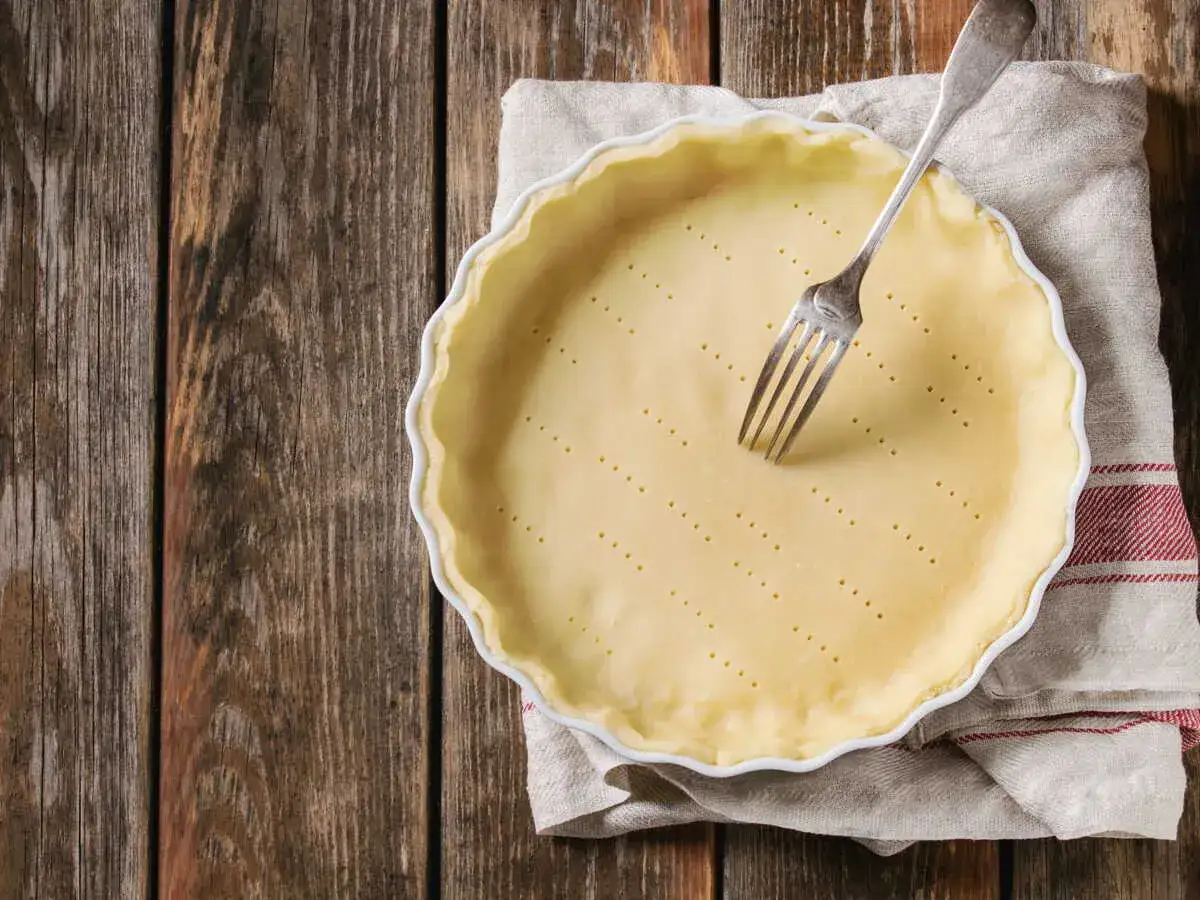 P&acirc;te bris&eacute;e sans beurre pr&ecirc;te &agrave; &ecirc;tre garnie, piqu&eacute;e &agrave; la fourchette dans un plat &agrave; tarte blanc sur un tissu &agrave; carreaux.