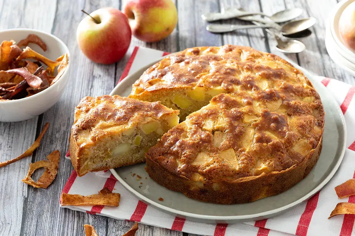 Un délicieux cake aux pommes, doré et moelleux, avec des morceaux de fruits visibles. Deux pommes fraîches et des chips de pomme complètent cette gourmandise.