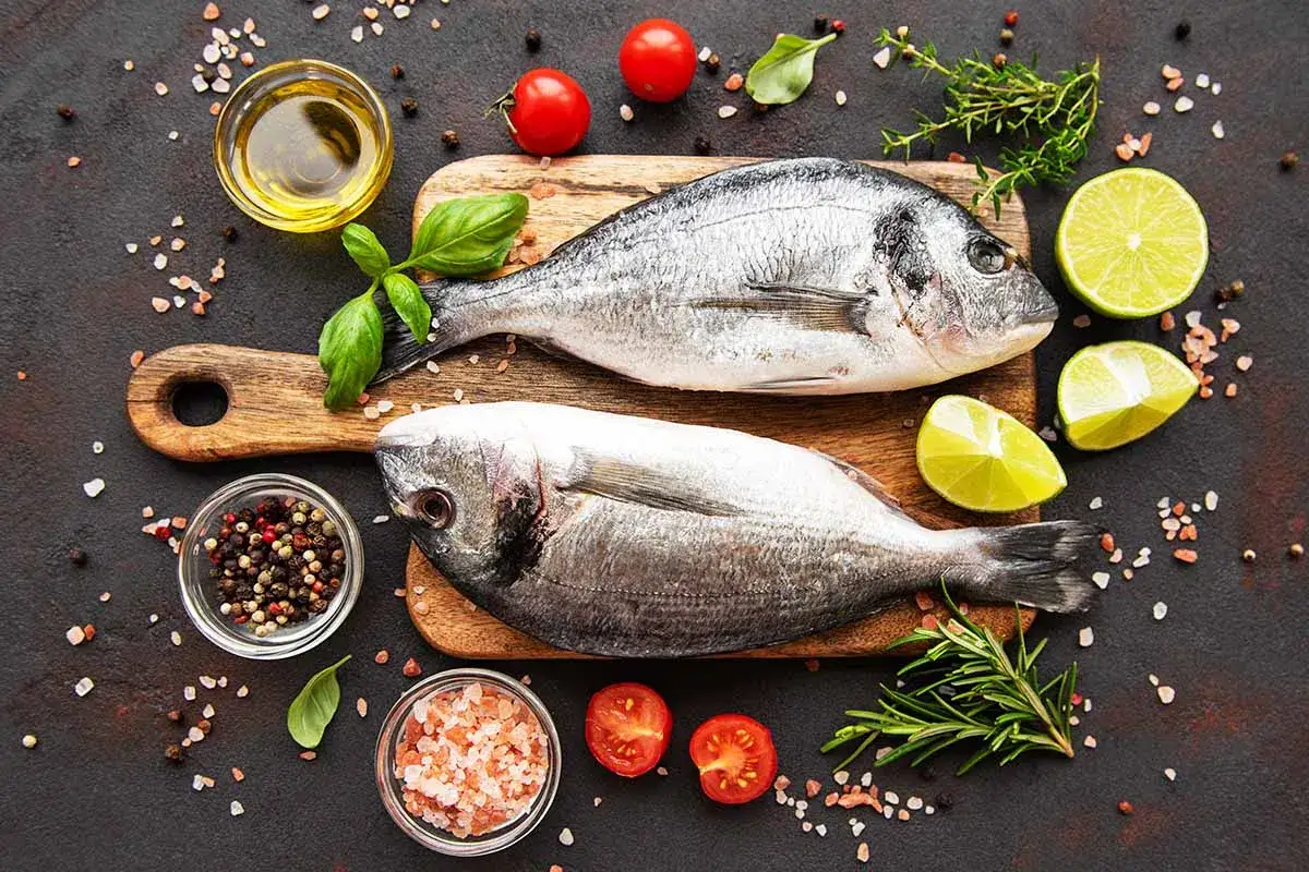 Deux poissons entiers pr&ecirc;ts pour la cuisson, accompagn&eacute;s de citrons, tomates, herbes et &eacute;pices.