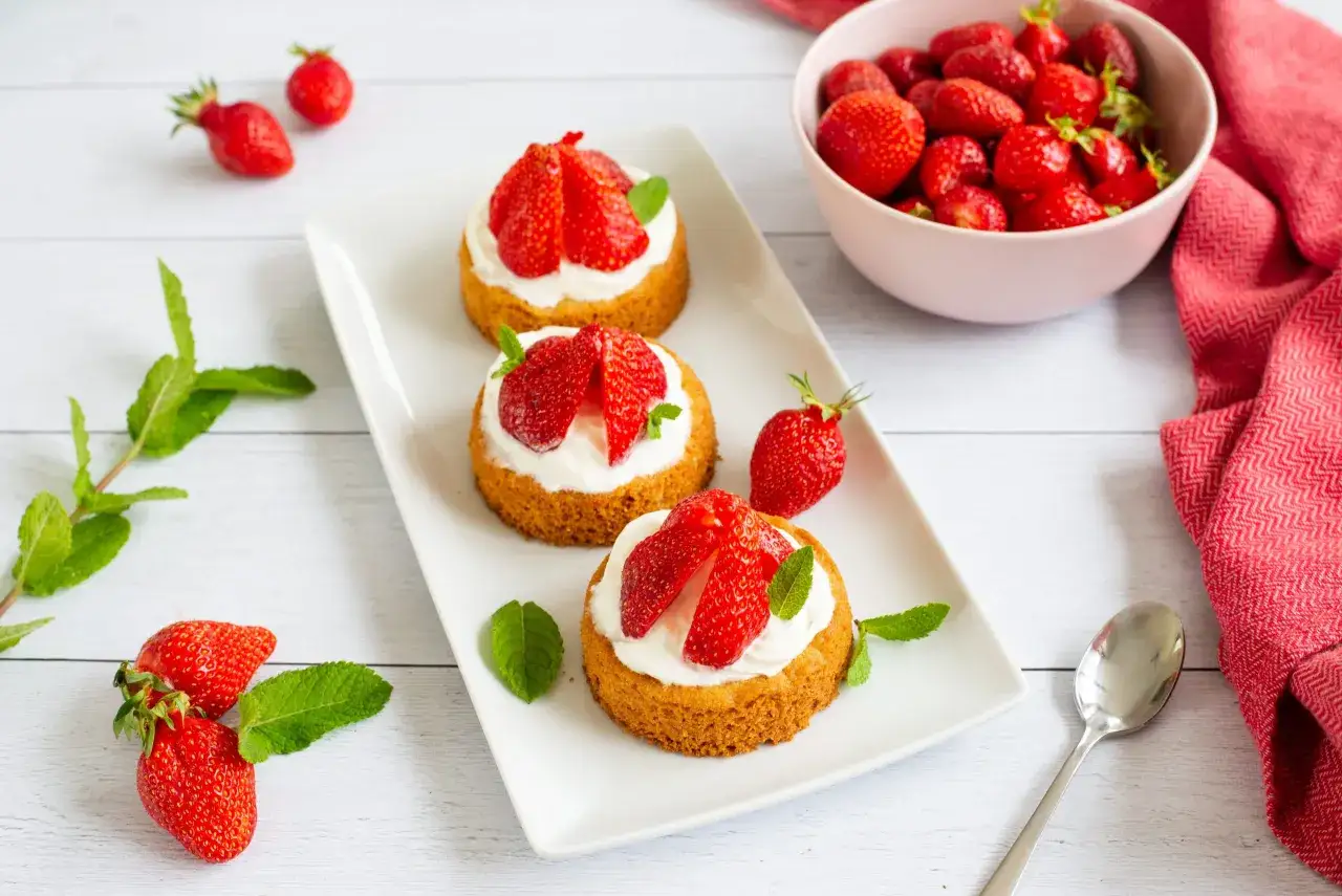 Trois petits g&acirc;teaux aux fraises, garnis de cr&egrave;me fouett&eacute;e et de fraises fra&icirc;ches, sur une assiette blanche.