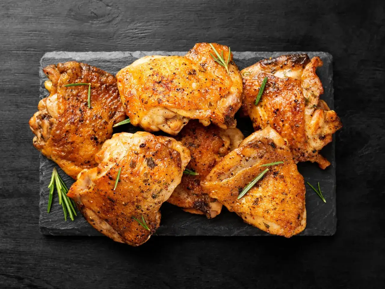 Plusieurs hauts de cuisse de poulet dor&eacute;s et croustillants, assaisonn&eacute;s d'herbes, sont dispos&eacute;s sur une planche d'ardoise.