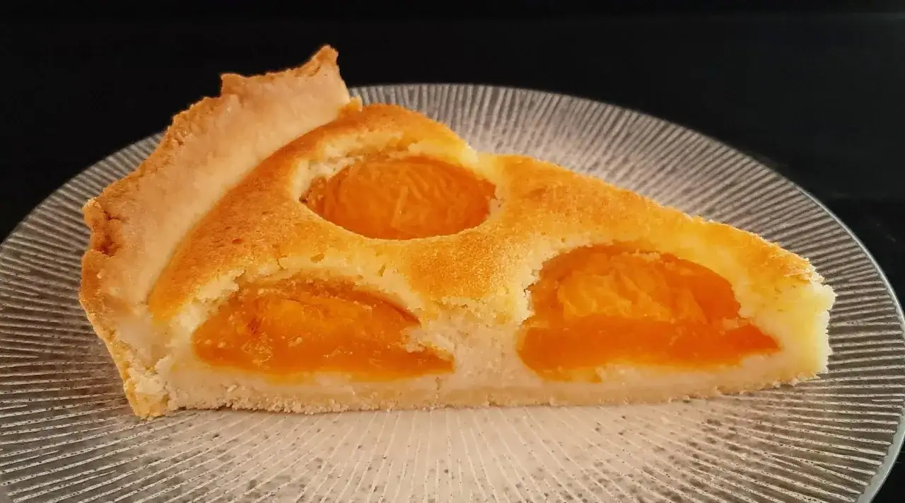 Une part de tarte aux abricots dorée, avec des fruits juteux incrustés dans une pâte moelleuse, sur une assiette texturée.