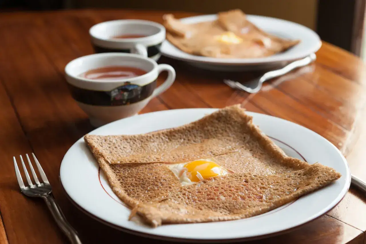 Une galette de sarrasin, sp&eacute;cialit&eacute; de la cr&ecirc;pe Bretagne, avec un &oelig;uf au centre, servie sur une assiette blanche avec une tasse de th&eacute;.