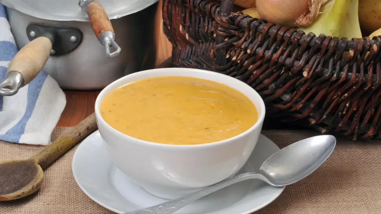 Un bol de soupe de l&eacute;gumes onctueuse, pr&ecirc;t &agrave; &ecirc;tre d&eacute;gust&eacute;. Une cuill&egrave;re en bois et un panier de l&eacute;gumes frais compl&egrave;tent cette sc&egrave;ne r&eacute;confortante.