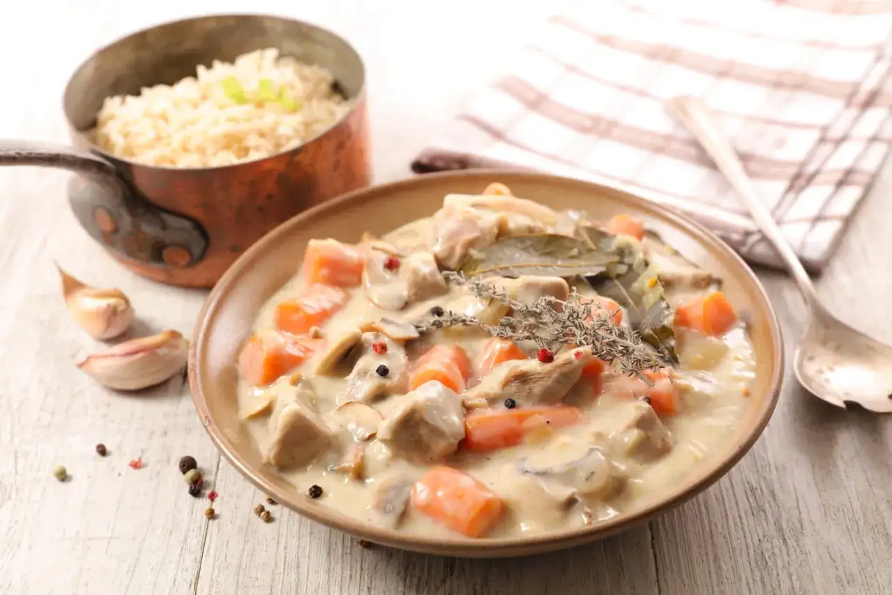 Un plat de blanquette de veau crémeuse avec des carottes, des champignons et des herbes, servi avec du riz dans une casserole en cuivre.