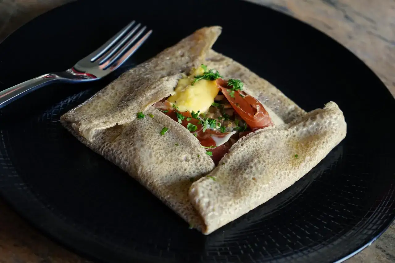 Cr&ecirc;pe garnie de jambon, fromage et herbes, sur une assiette noire. Un clin d'&oelig;il &agrave; l'origine des cr&ecirc;pes sucr&eacute;es.