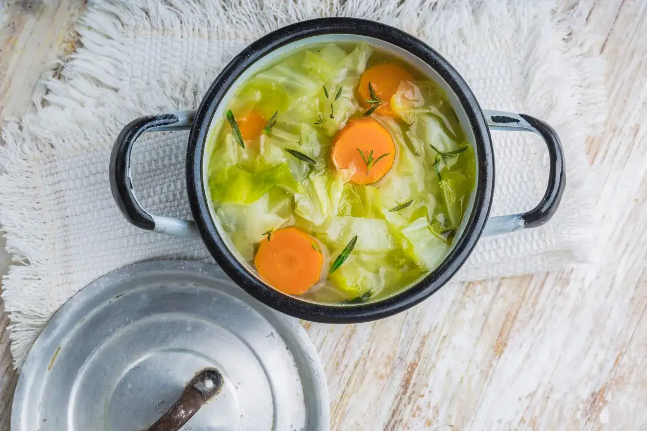 Une marmite de soupe aux choux, avec des carottes et des herbes, sur une serviette blanche.