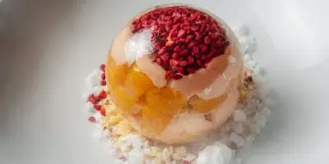 Sphère transparente garnie de fruits rouges et de pêche melba, sur un lit de miettes et de glace pilée.
