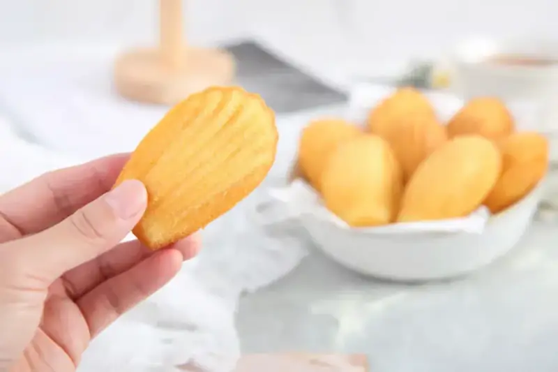 Une main tient une madeleine dorée, parfaite pour une recette madeleine moelleuse. Un bol rempli de ces délicieuses pâtisseries est visible en arrière-plan.