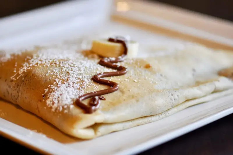 Crêpe pliée, saupoudrée de sucre glace, décorée de chocolat fondu et d'une rondelle de banane. La meilleure recette de pâte à crêpes pour un délice !